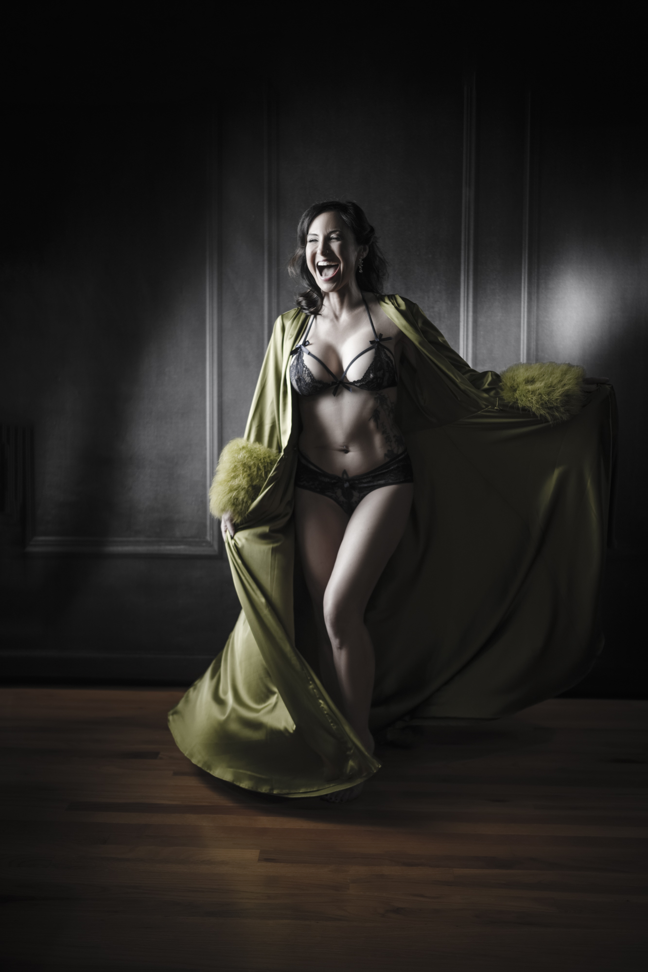 A woman laughing joyfully in a black lace set and flowing olive robe under dramatic low-key studio lighting